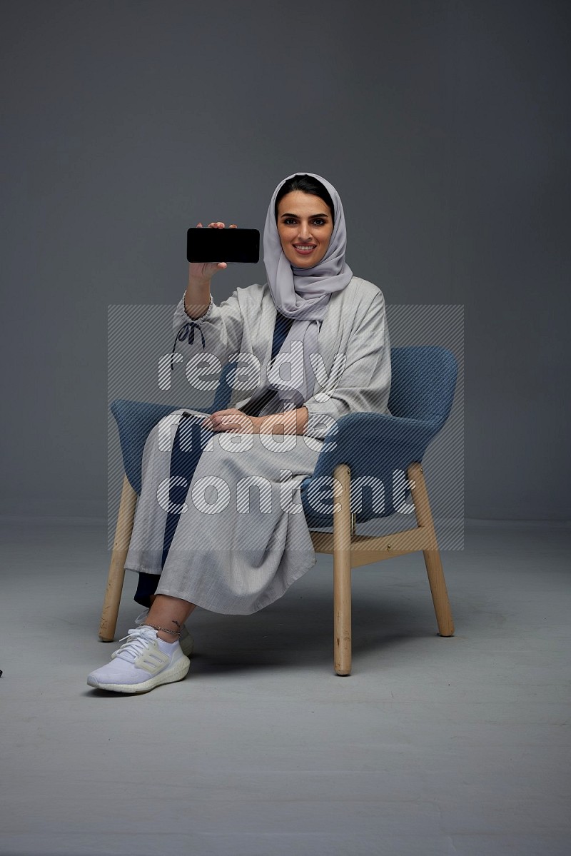 A Saudi woman wearing a light gray Abaya and head scarf sitting on a dark grey chair and showing the phone's screen eye level on a grey background