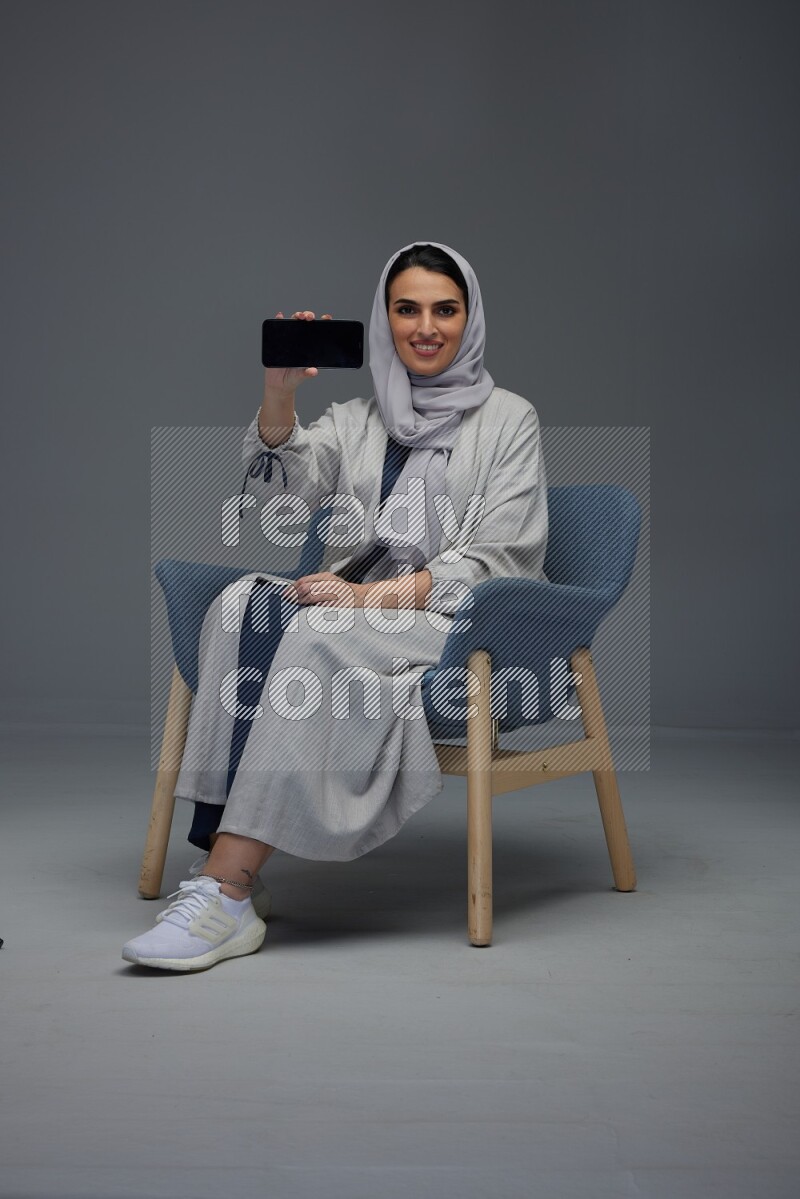 A Saudi woman wearing a light gray Abaya and head scarf sitting on a dark grey chair and showing the phone's screen eye level on a grey background