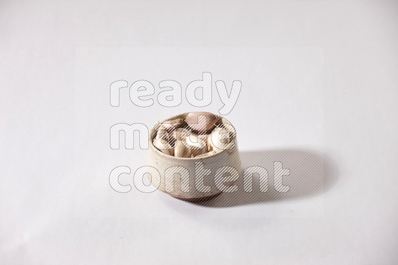 A beige pottery bowl full of garlic cloves on a white flooring in different angles