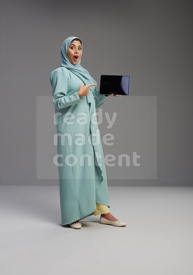 Saudi Woman wearing Abaya standing showing tablet to camera on Gray background