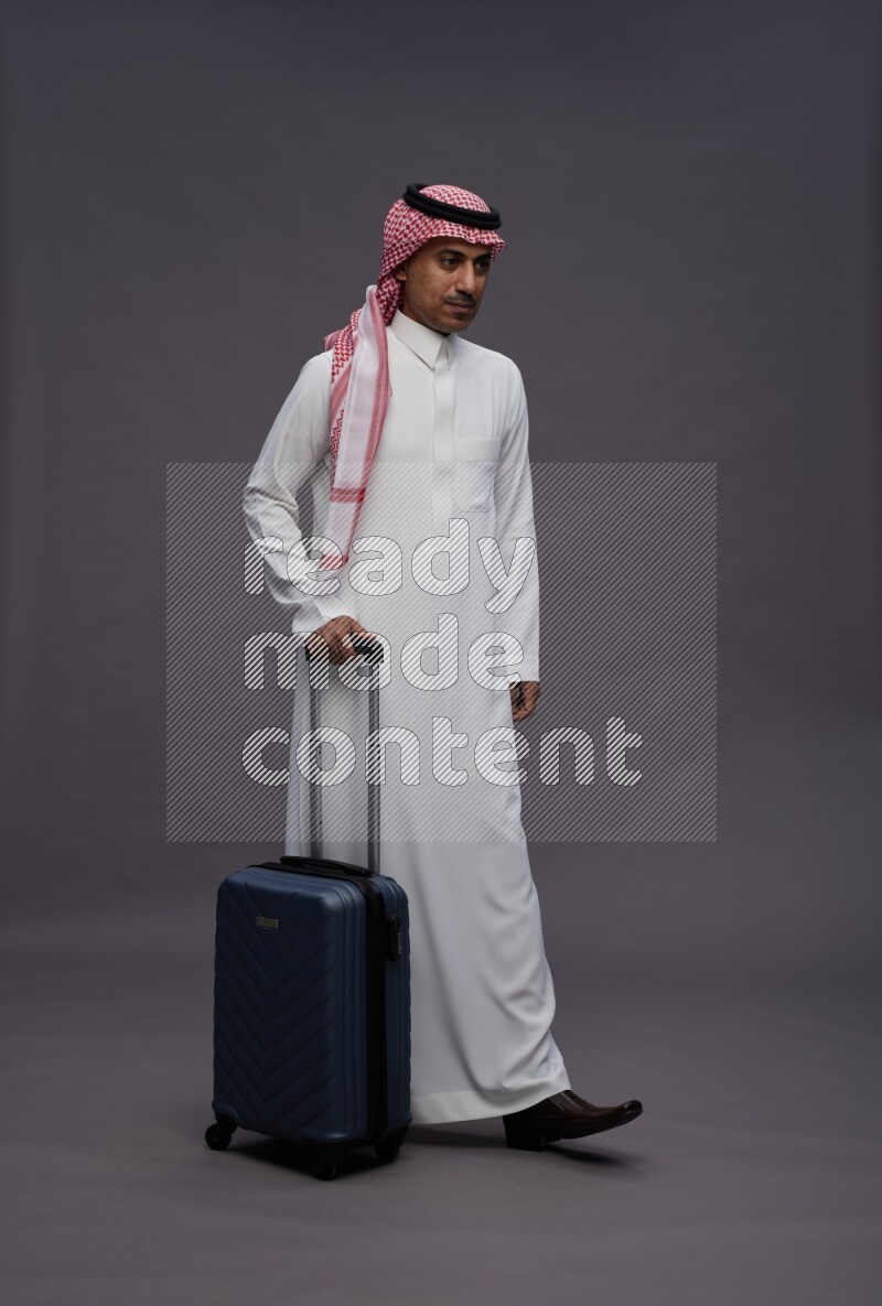 Saudi man wearing thob and shomag standing holding bag on gray background