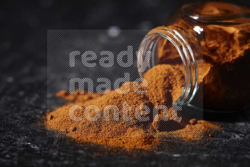 A glass jar full of ground paprika powder flipped with some spilling powder on black background