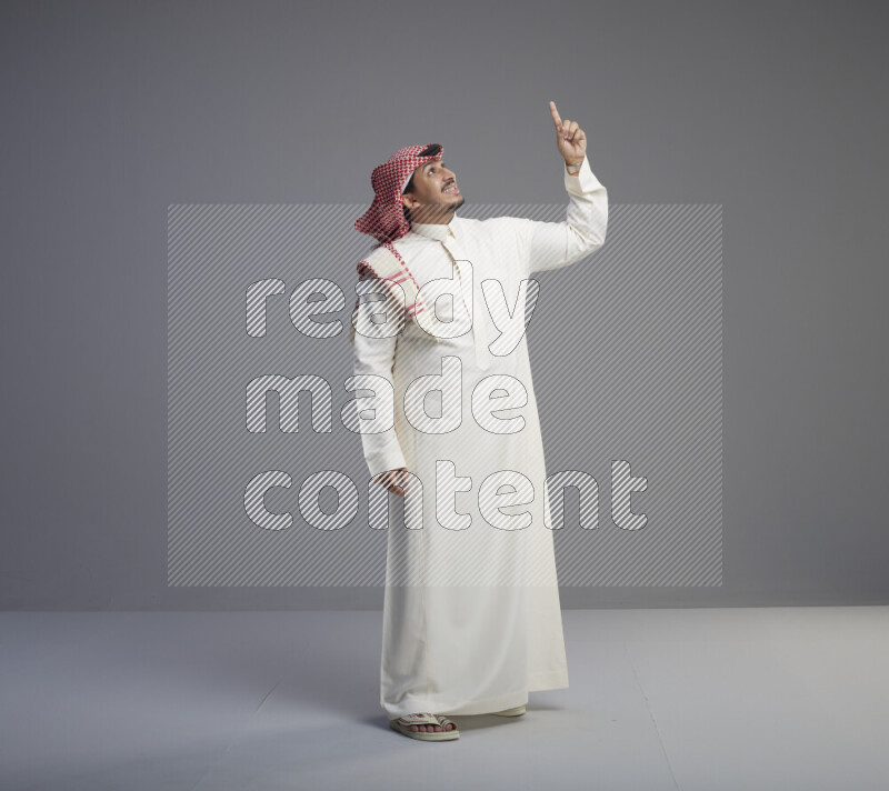 A Saudi man standing wearing thob and red shomag interacting with the camera on gray background