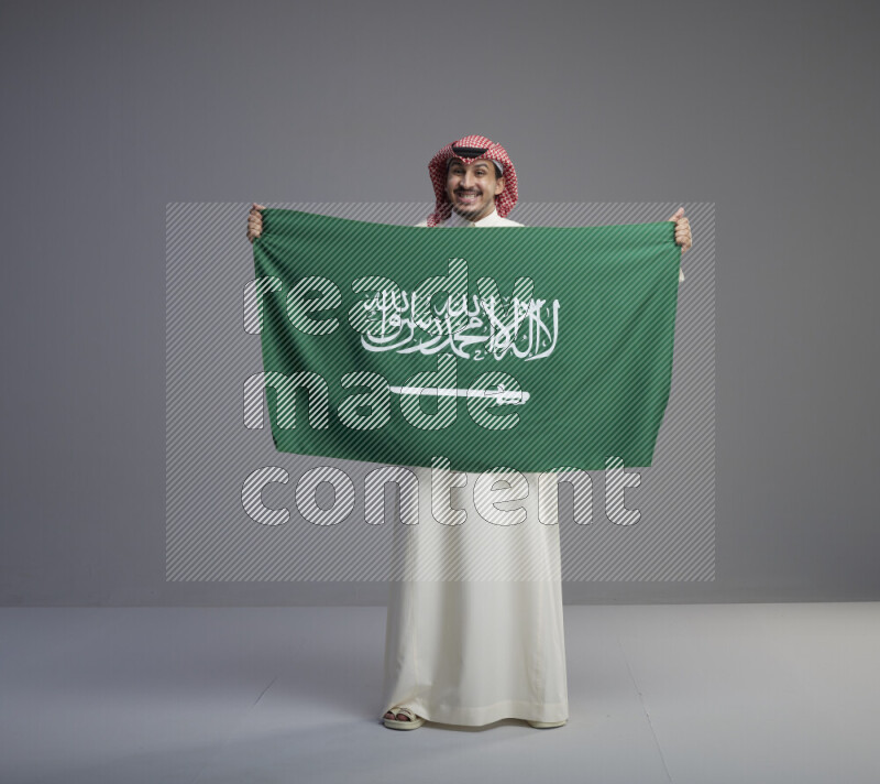 A saudi man standing wearing thob and red shomag holding big saudi flag on gray background