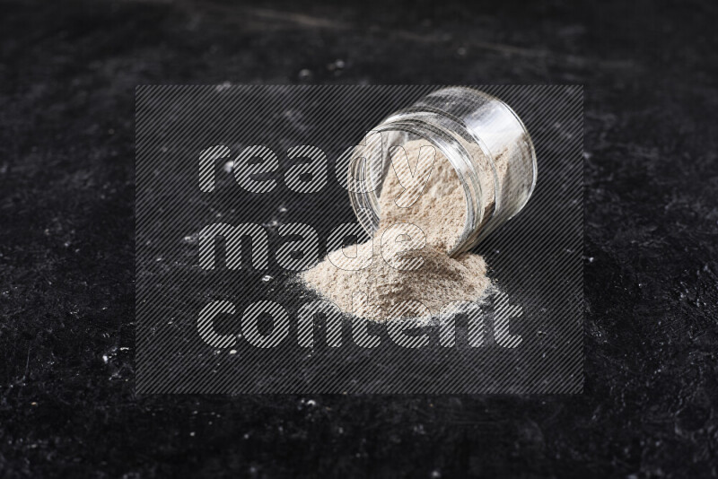 A glass jar full of onion powder flipped with some spilling powder on black background