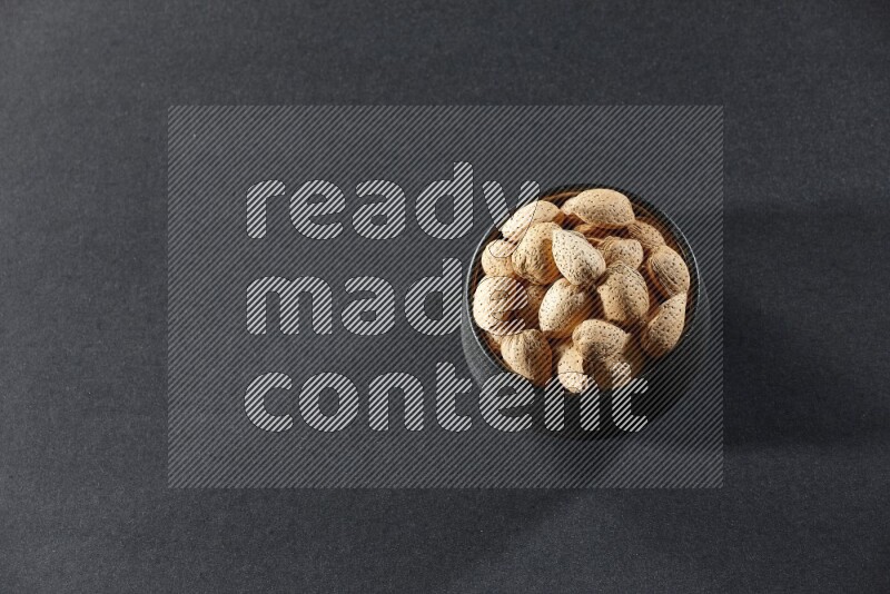 A black pottery bowl full of almonds on a black background in different angles