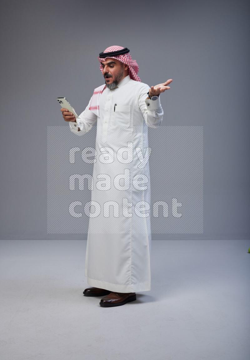 Saudi man Wearing Thob and red Shomag standing texting on phone on Gray background