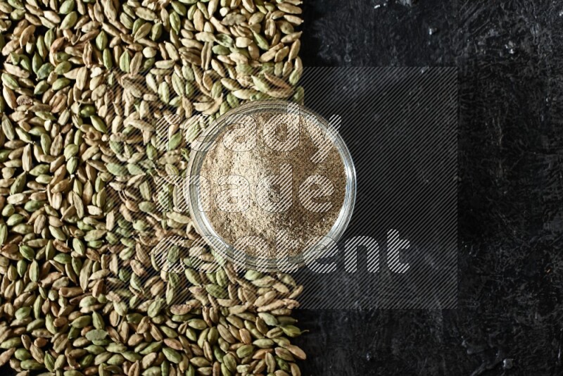 A glass bowl full of cardamom powder and cardamom seeds spreaded beneath the bowl on textured black flooring