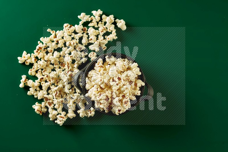 A black ceramic bowl full of popcorn with popcorn beside it on a green background in a top view shot