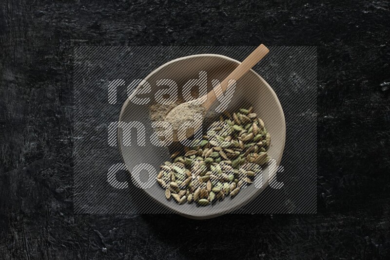 A plate filled with cardamom seeds and a wooden spoon full of cardamom powder on a textured black flooring