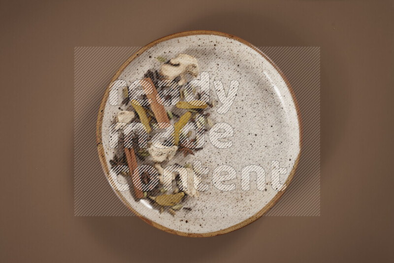 An assorted whole spices on a flat beige plate including fresh and dried ginger, cinnamon sticks, cardamom, turmeric fingers, cloves, star anise on a beige background