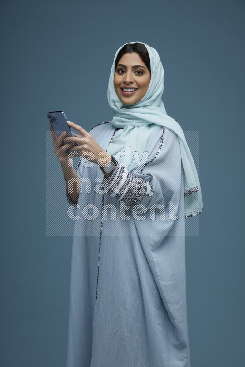 A Saudi woman posing with her phone in a blue background wearing a blue Abaya with hijab