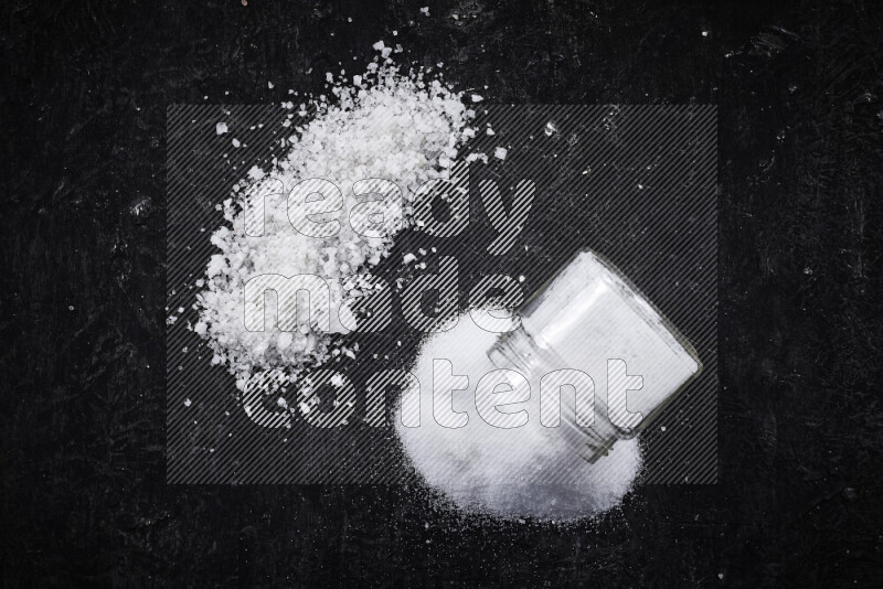 A glass jar full of table salt with some sea salt crystals beside it on a black background