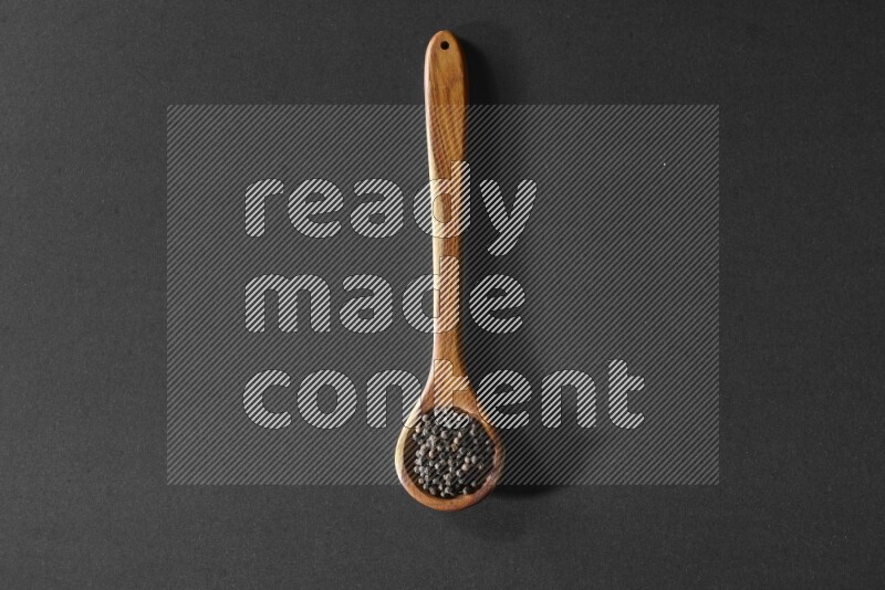 A wooden ladle full of black pepper beads on a black flooring