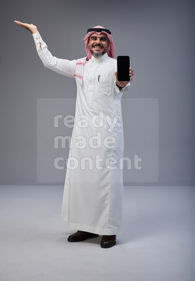 Saudi man Wearing Thob and red Shomag standing showing phone to camera on Gray background