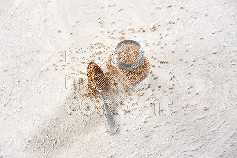 A glass spice jar and a metal spoon full of mustard seeds on a textured white flooring