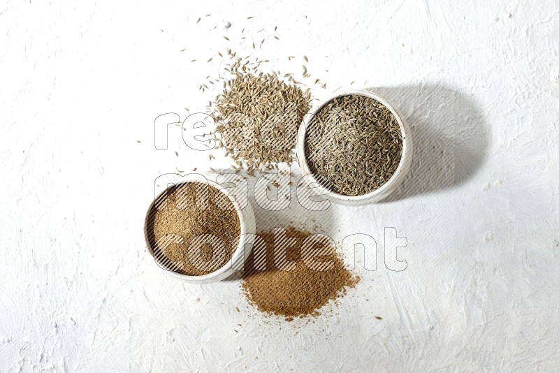 2 beige bowls full of cumin seeds and powder with spilled powder and seeds on textured white flooring