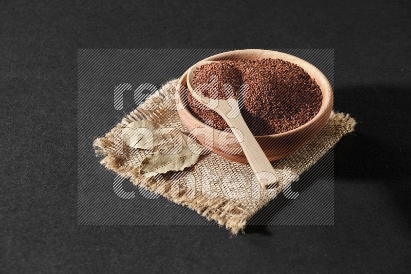 A wooden bowl full of garden cress seeds with wooden spoon full of the seeds on it on burlap fabric on a black flooring