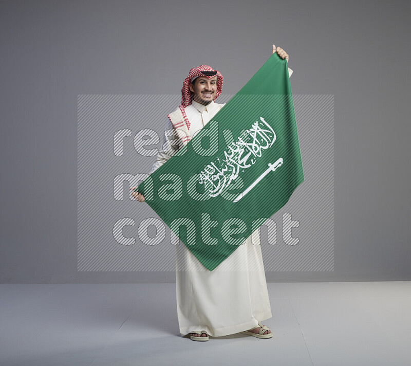 رجل سعودي يرتدي ثوب ابيض وشماغ احمر ويحمل علم كبير للسعودية