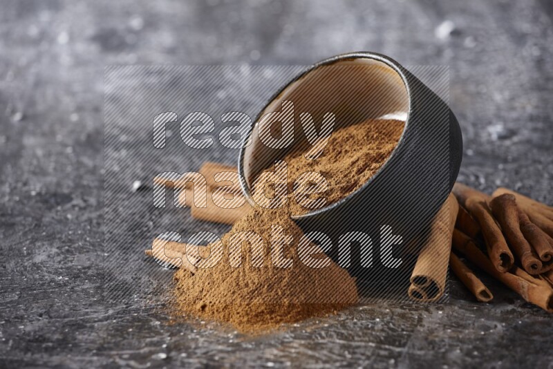 Black pottery bowl over filled with cinnamon powder and cinnamon sticks around the bowl on a textured black background