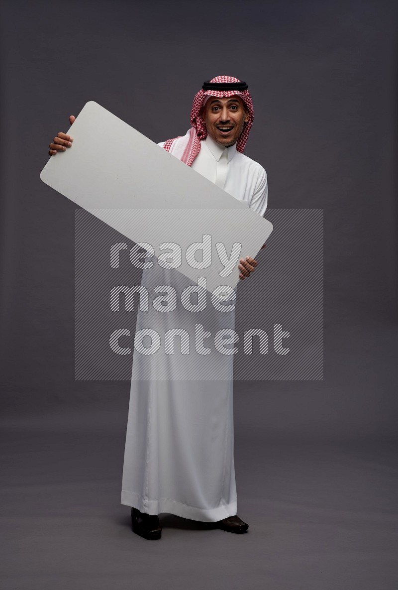 Saudi man wearing thob and shomag standing holding board on gray background