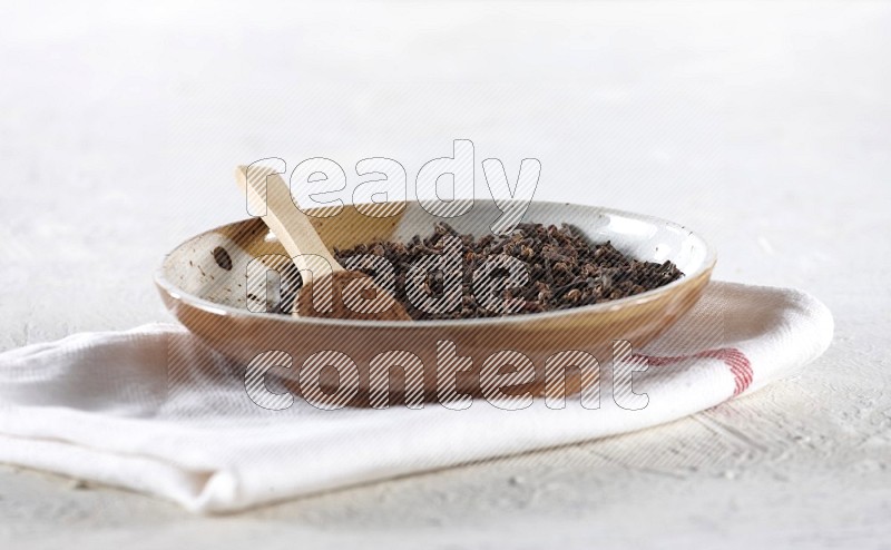 A Pottery plate full of cloves and a on it a wooden spoon full of cloves powder on a textured white background