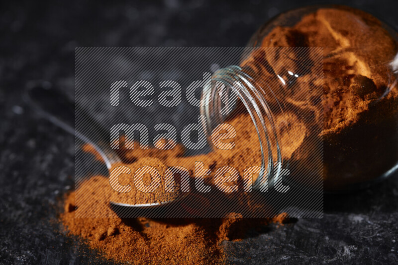 A glass jar full of ground paprika powder flipped with some spilling powder on black background