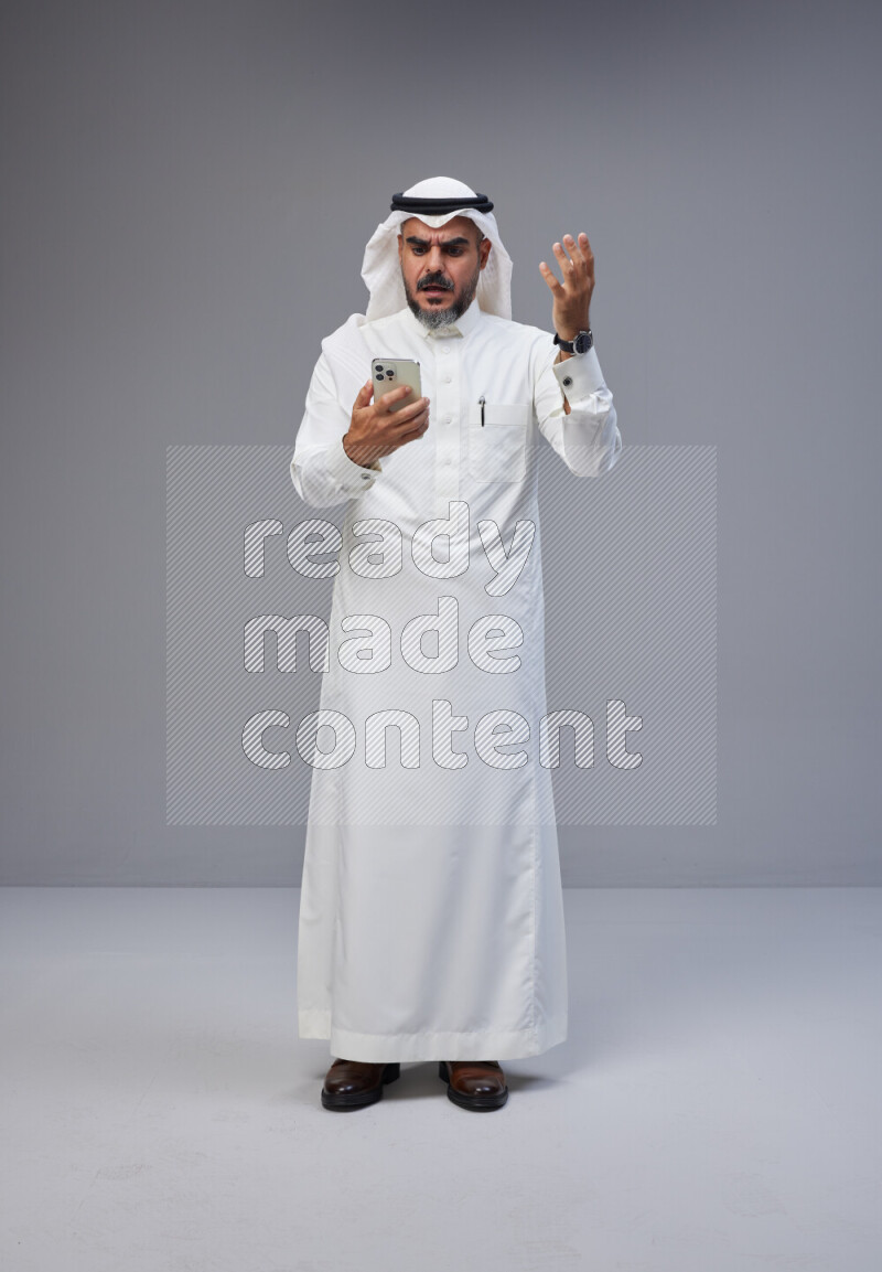 Saudi man Wearing Thob and white Shomag standing texting on phone on Gray background
