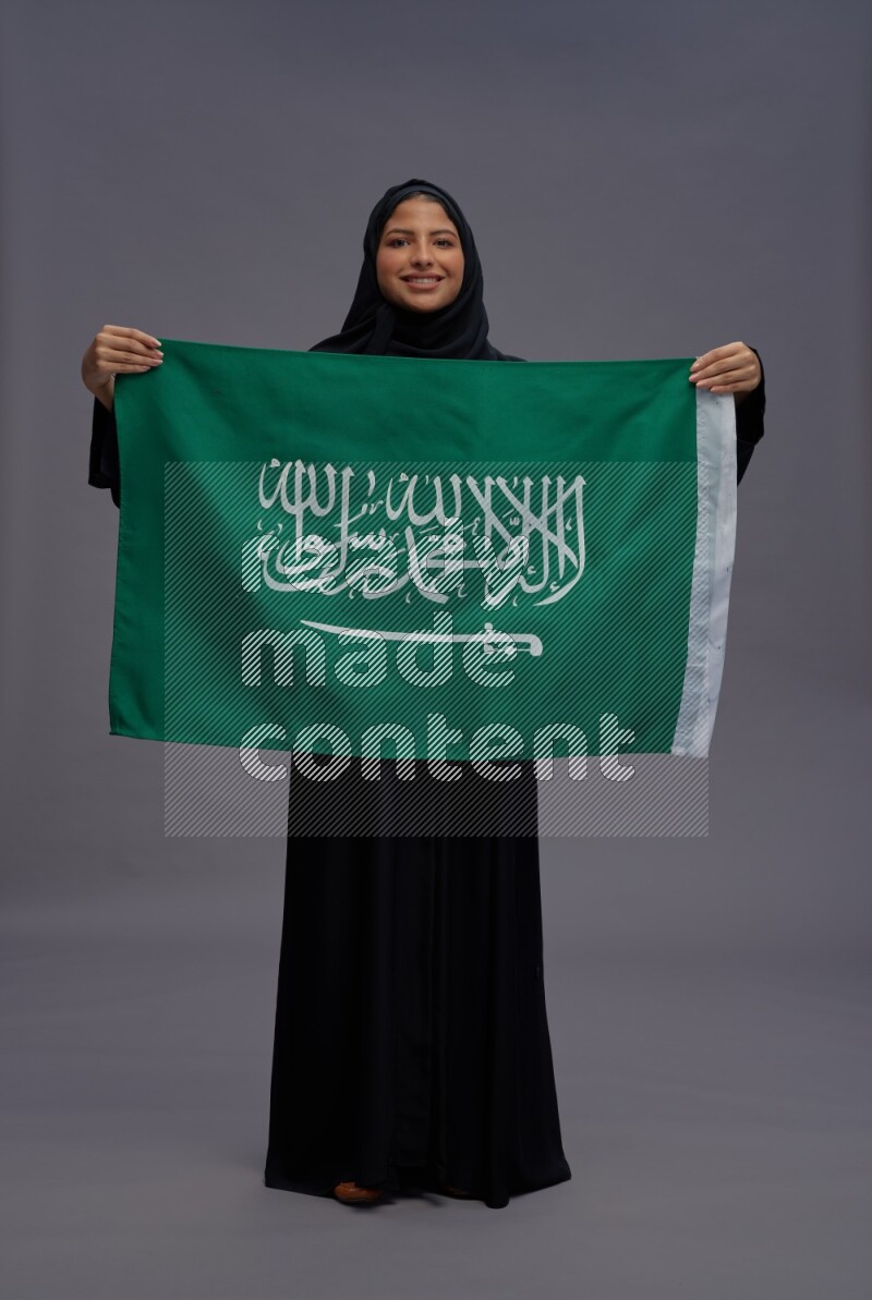 Saudi woman wearing Abaya standing holding Saudi flag on gray background