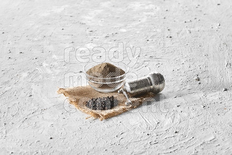 A glass bowl full of black pepper powder, black pepper beads and a turkish metal grinder on burlap fabric on textured white flooring