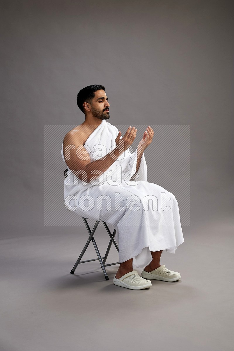 A man wearing Ehram sitting on chair performing dua'a on gray background