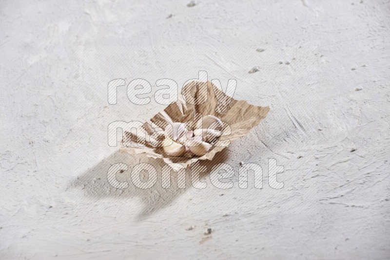 A crumpled piece of paper full of garlic cloves on a textured white flooring in different angles