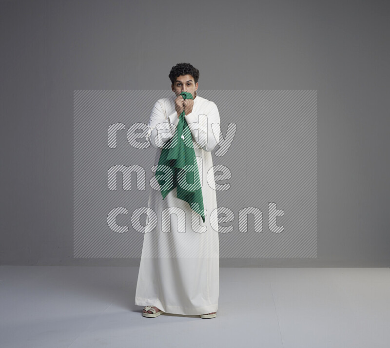 A Saudi man standing wearing thob kissing Saudi flag on gray background