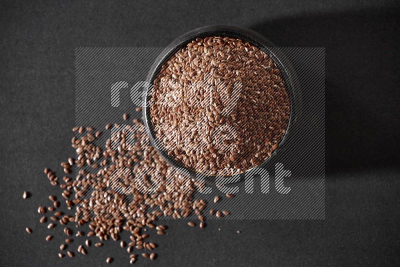 A black pottery bowl full of flaxseeds surrounded by the seeds on a black flooring