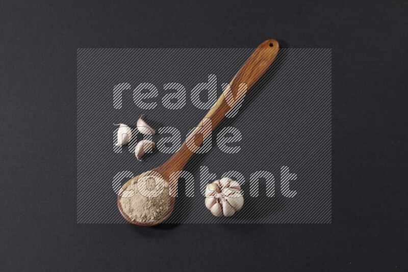 A wooden ladle full of garlic powder with a whole garlic bulb and some cloves beside it on a black flooring