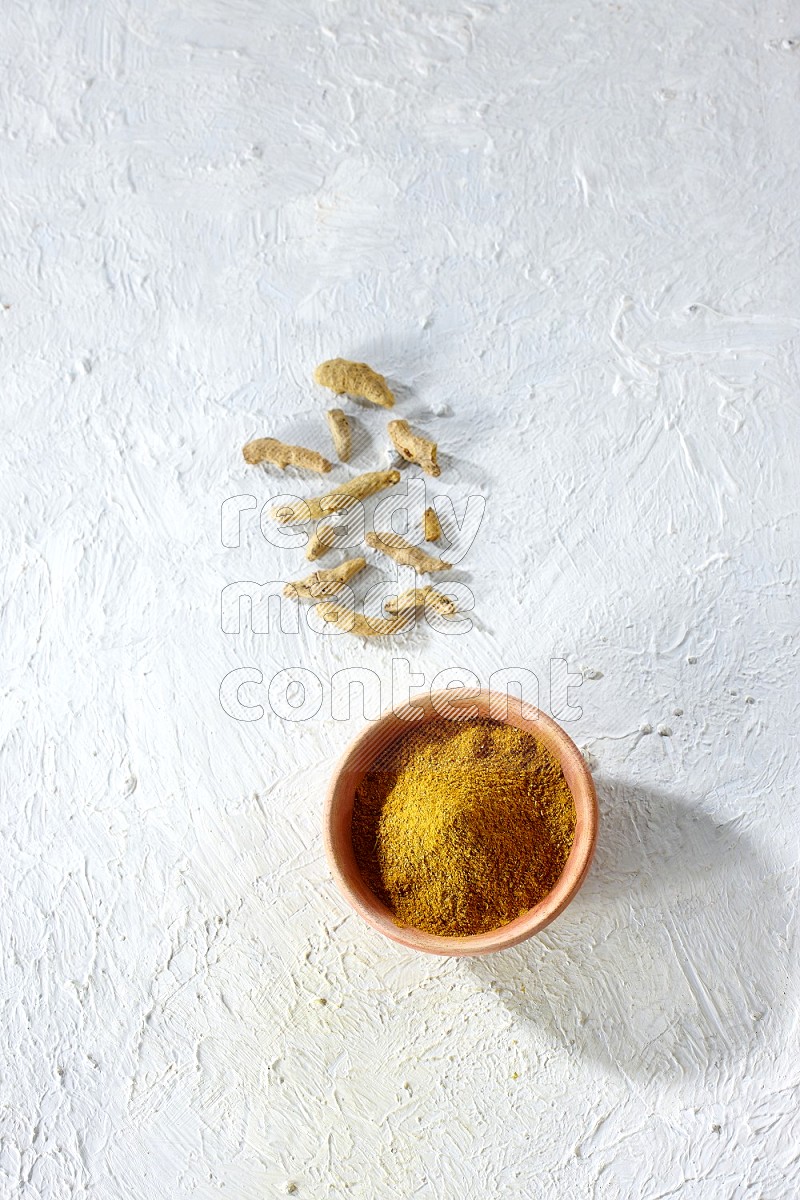 A wooden bowl full of turmeric powder with dried whole fingers on textured white flooring