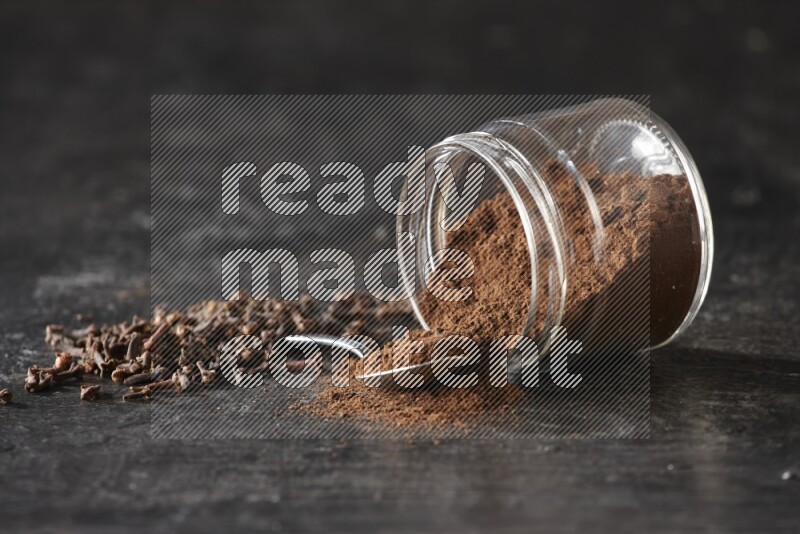 A flipped glass jar and metal spoon full of cloves powder with cloves spread on a textured black flooring