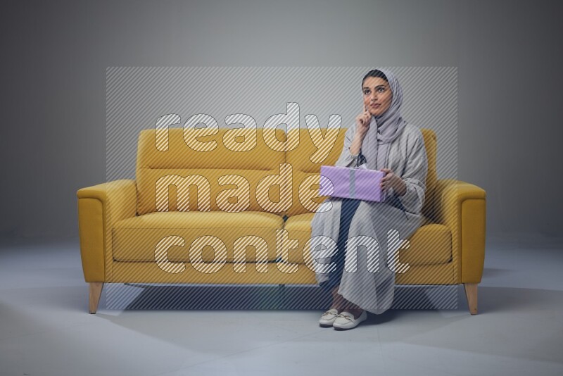 A Saudi woman wearing a light gray Abaya and head scarf sitting on a yellow sofa holding a gift box beside multi color gift boxes eye level on a grey background
