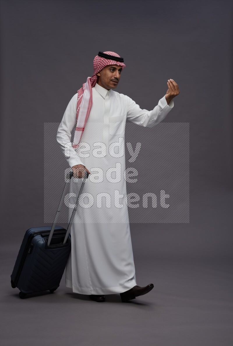 Saudi man wearing thob and shomag standing holding bag on gray background