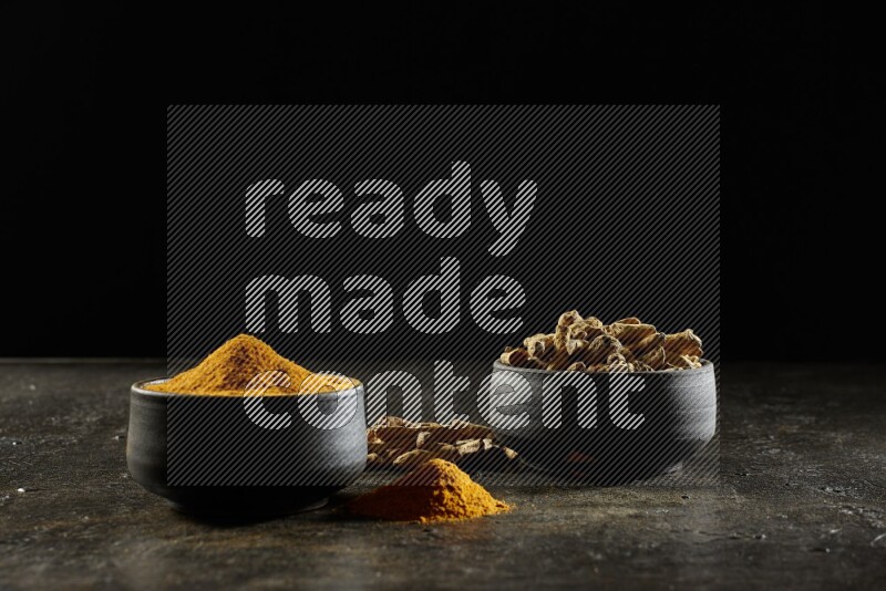 2 pottery black bowls, one full of turmeric powder and the other full of dried turmeric whole fingers on a textured black flooring
