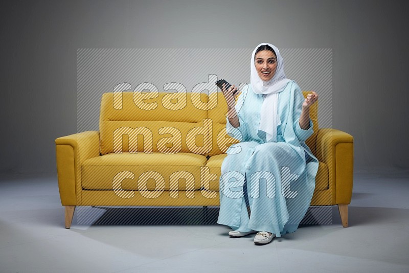 A Saudi woman wearing a light blue Abaya and white head scarf sitting on a yellow sofa and texting on her phone on a grey background