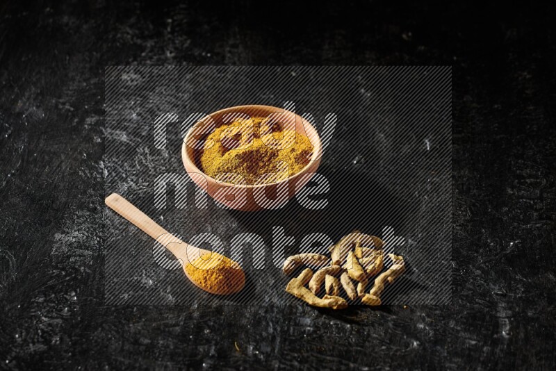 A wooden bowl and a wooden spoon full of turmeric powder with dried turmeric fingers on textured black flooring