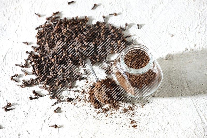 A glass spice jar and a metal spoon full of cloves powder and cloves spread on textured white flooring
