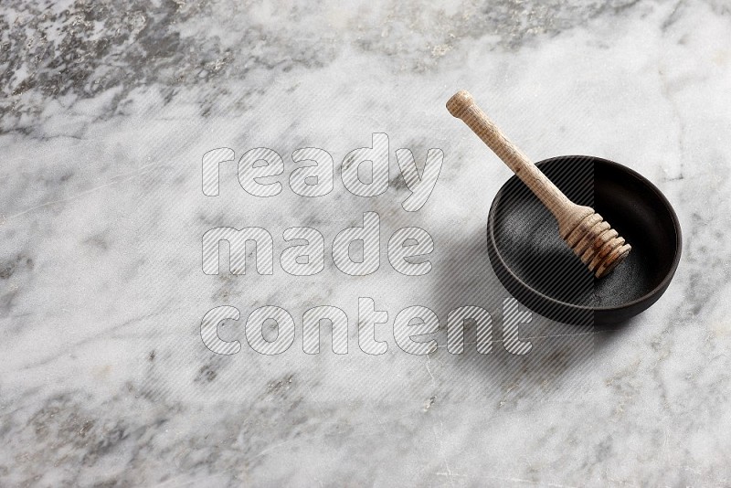 Black Pottery Bowl with wooden honey handle in it, on grey marble flooring, 65 degree angle