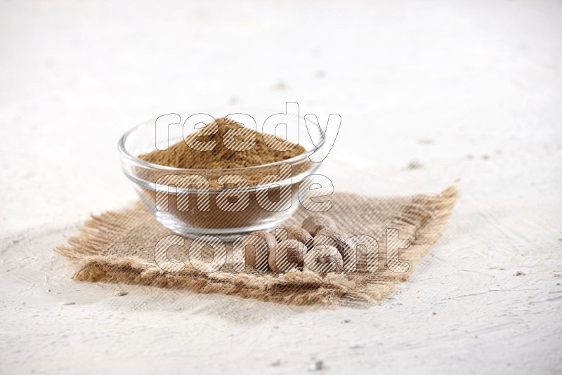 A glass bowl full of nutmeg powder with the seeds beside it on burlap fabric on a textured white flooring in different angles