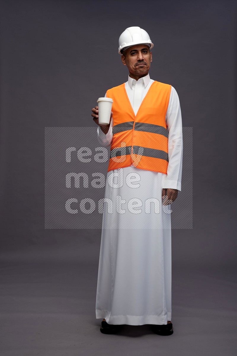 Saudi man wearing thob with engineer vest standing holding paper cup on gray background