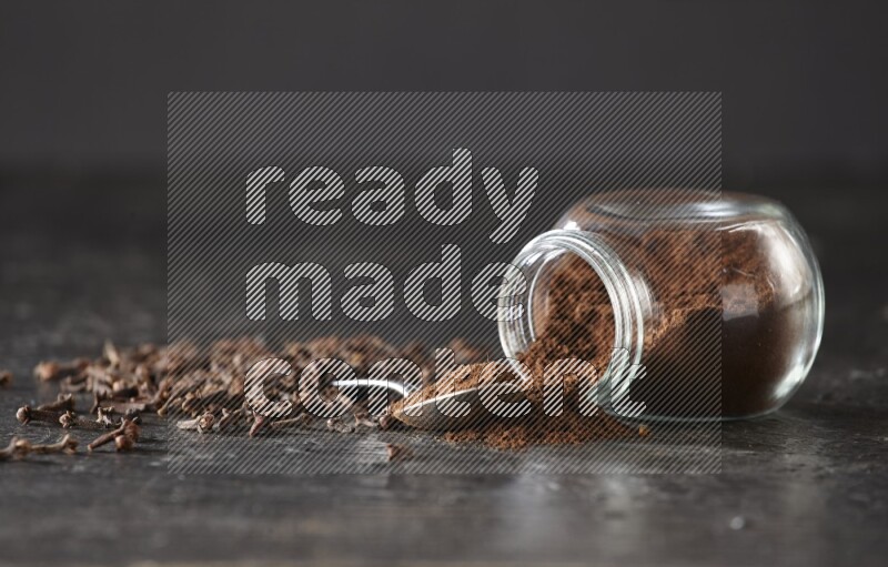 A flipped glass spice jar and a metal spoon full of cloves powder with cloves spread on textured black flooring