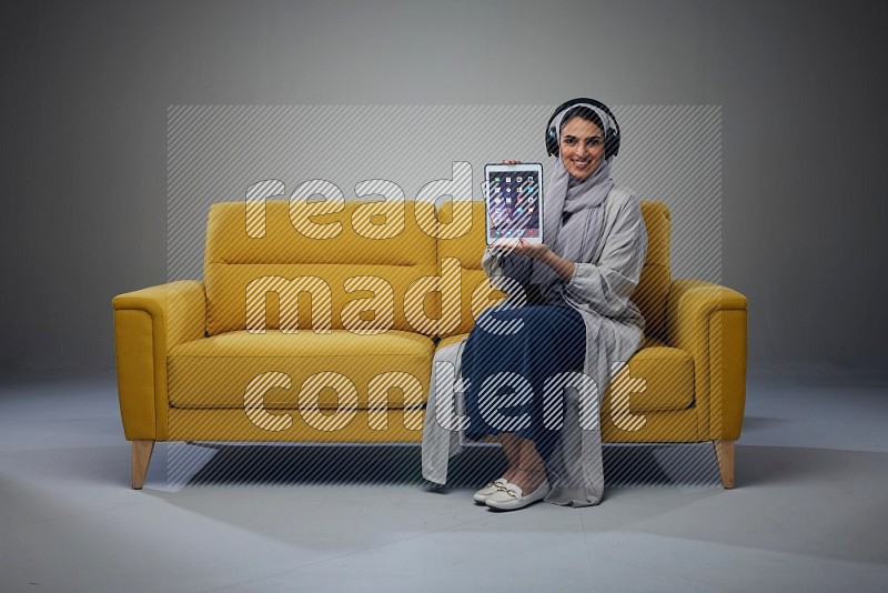 A Saudi woman wearing a light gray Abaya and head scarf sitting on a yellow sofa and using her phone while wearing headphones eye level on a grey background