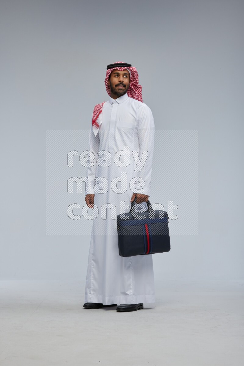 Saudi man Wearing Thob and shomag standing holding bag on Gray background