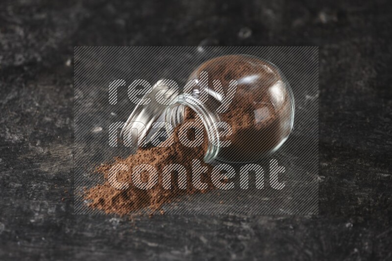 A flipped glass spice jar full of cloves powder and powder came out of it on textured black flooring